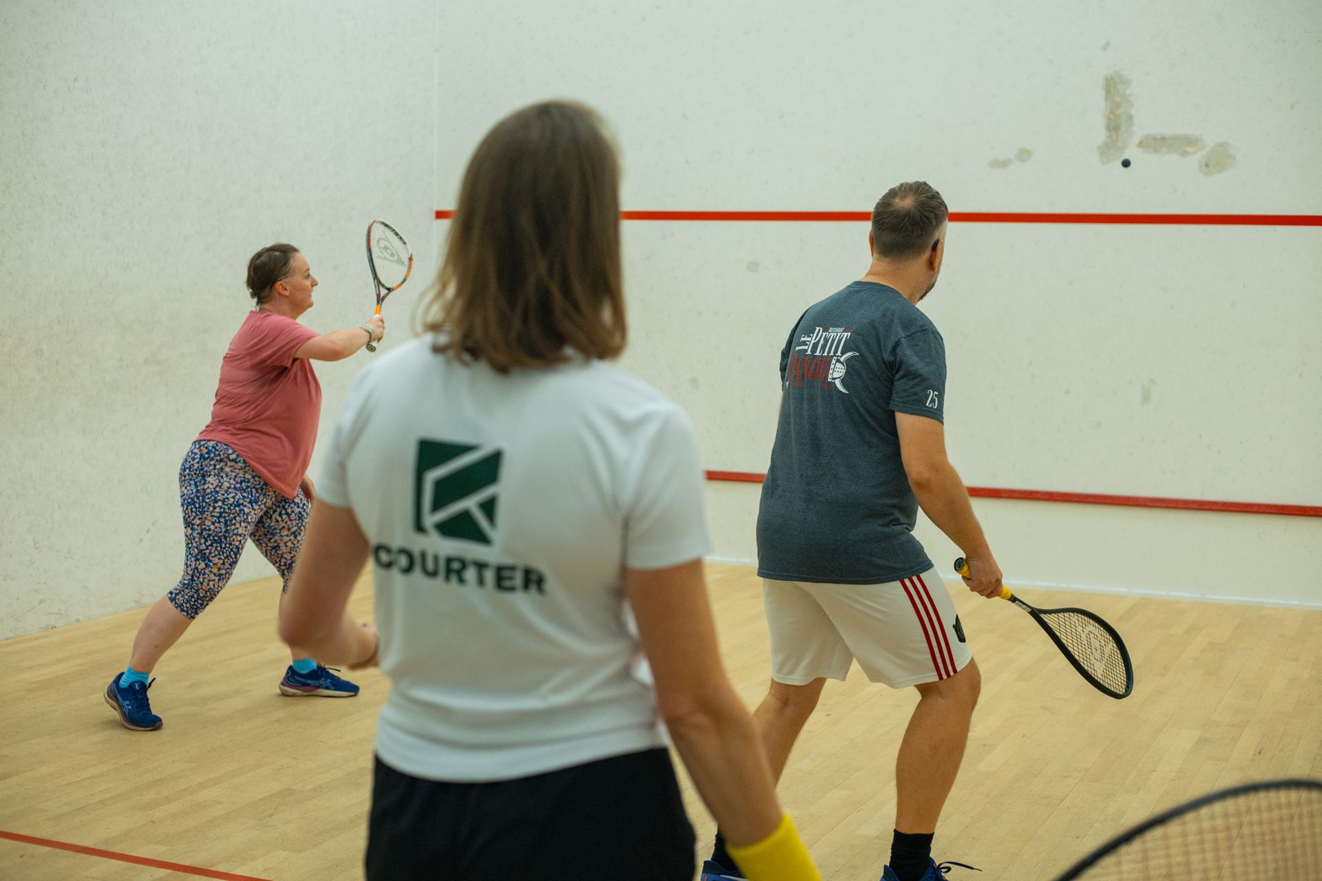 A couple rallying on a squash court, watched by their coach in a Courter T-shirt