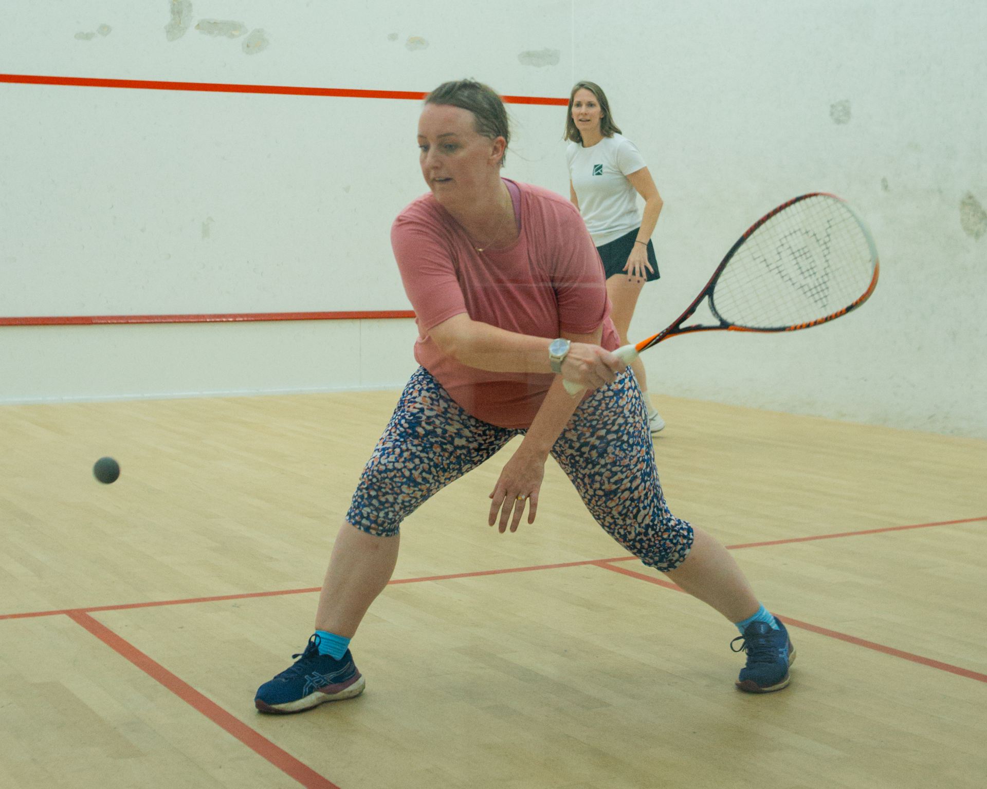 A woman lunging for a low ball on a squash court as her coach watches