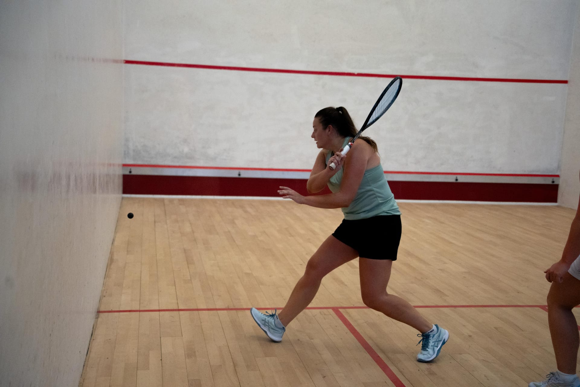 A coach in a mint Courter vest stretching for the ball near the front of a squash court