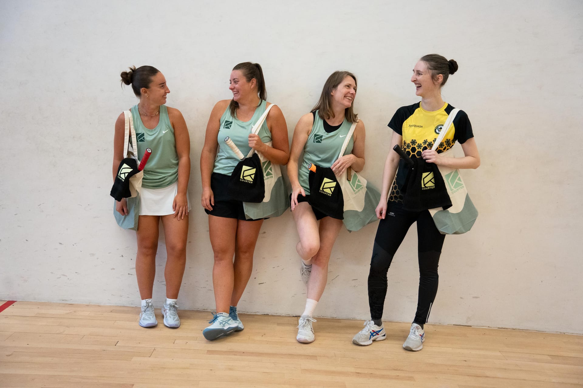 Four Courter coaches leaning on a court wall with tote bags over their shoulders