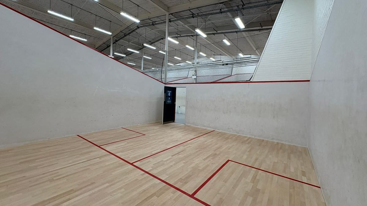 An empty squash court with a pale wooden floor and red lines, viewed from the back of the court