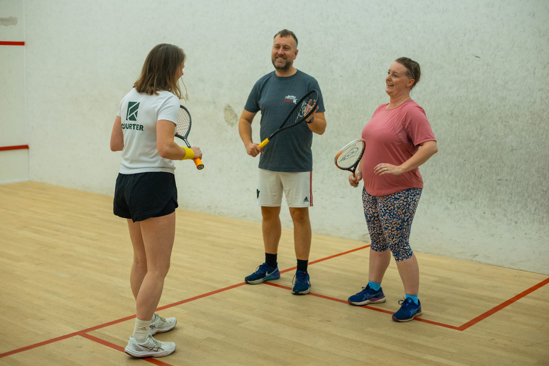 A coach in a white Courter T-shirt laughing with a couple on a squash court